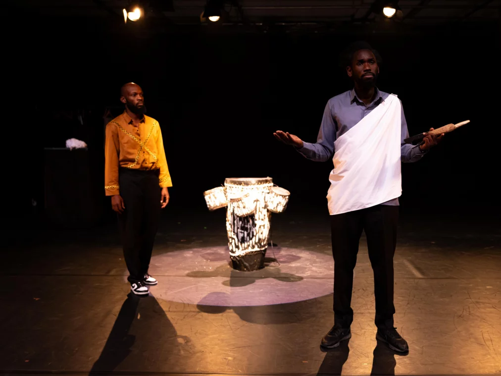 Zwei Personen stehen auf einer dunklen Bühne: eine Person im goldgelben Hemd im Hintergrund, eine Person im hellen Hemd mit weißem Tuch über der Schulter im Vordergrund. Zwischen ihnen steht eine trommelartige Skulptur im Spotlicht, während die vordere Person mit offenen Händen spricht.