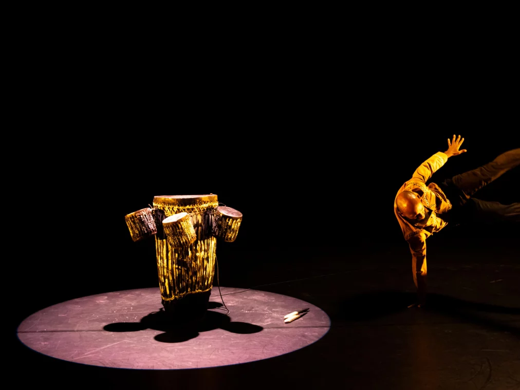 Eine Person im goldgelben Hemd führt einen akrobatischen Bodentrick aus und stützt sich auf eine Hand, während die Beine seitlich ausgestreckt in der Luft schweben. Links steht die trommelartige Skulptur im Spotlicht, der Hintergrund bleibt tiefschwarz.