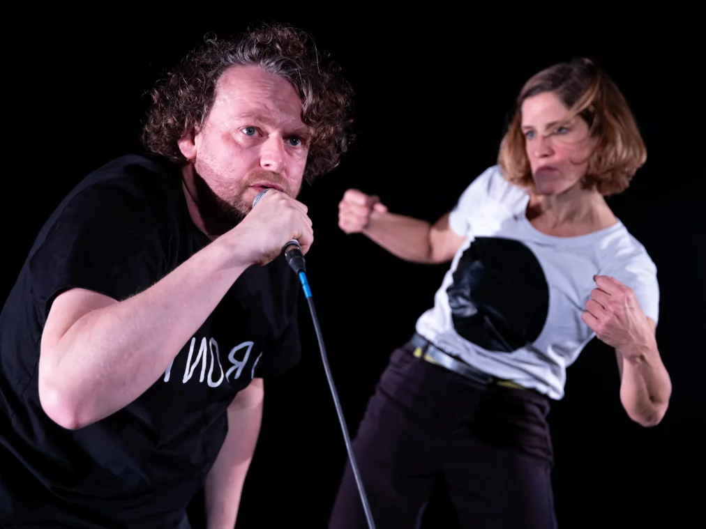 Zwei Personen auf dunkler Bühne: Im Vordergrund beugt sich eine Person mit Mikrofon intensiv nach vorn, während die Person im Hintergrund eine kraftvolle Kampfpose einnimmt, mit angespannten Armen und fokussiertem Blick.