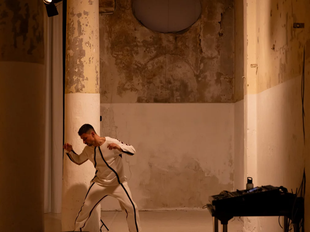 Eine Person in einem weißen Trainingsanzug steht in einer schmalen, hohen Raumnische vor einer verwitterten Wand und beugt sich dynamisch nach vorne, als würde sie in eine Bewegung hineinstarten.