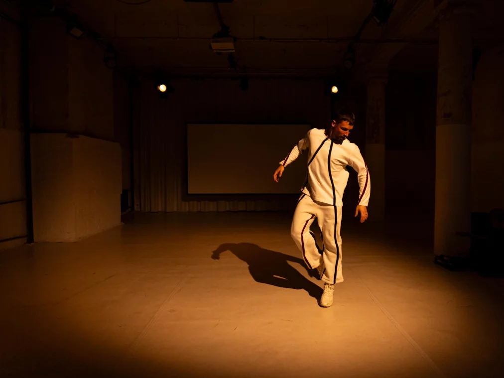 Eine Person in einem weißen Trainingsanzug bewegt sich tänzerisch über die leere Fläche des Raums, während im Hintergrund ein unbeleuchteter Projektionsschirm hängt. Warmes Licht wirft einen langen Schatten der Person über den Boden.
