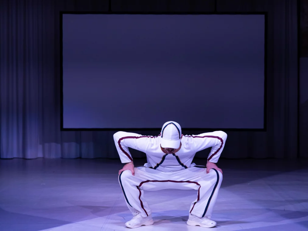 Eine Person in weißer Trainingskleidung hockt breitbeinig und tief am Boden, den Blick nach unten gerichtet. Der Körper bildet eine kraftvolle, stabile Haltung, während ein großer Projektionsschirm im Hintergrund zu sehen ist.