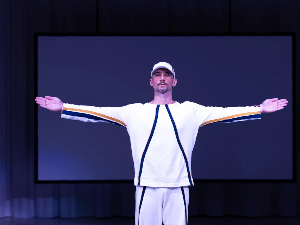 Eine Person im weißen Trainingsanzug steht frontal zum Publikum, die Arme weit ausgestreckt. Hinter ihr hängt ein großer dunkler Projektionsschirm. Das Licht ist kühl und klar.