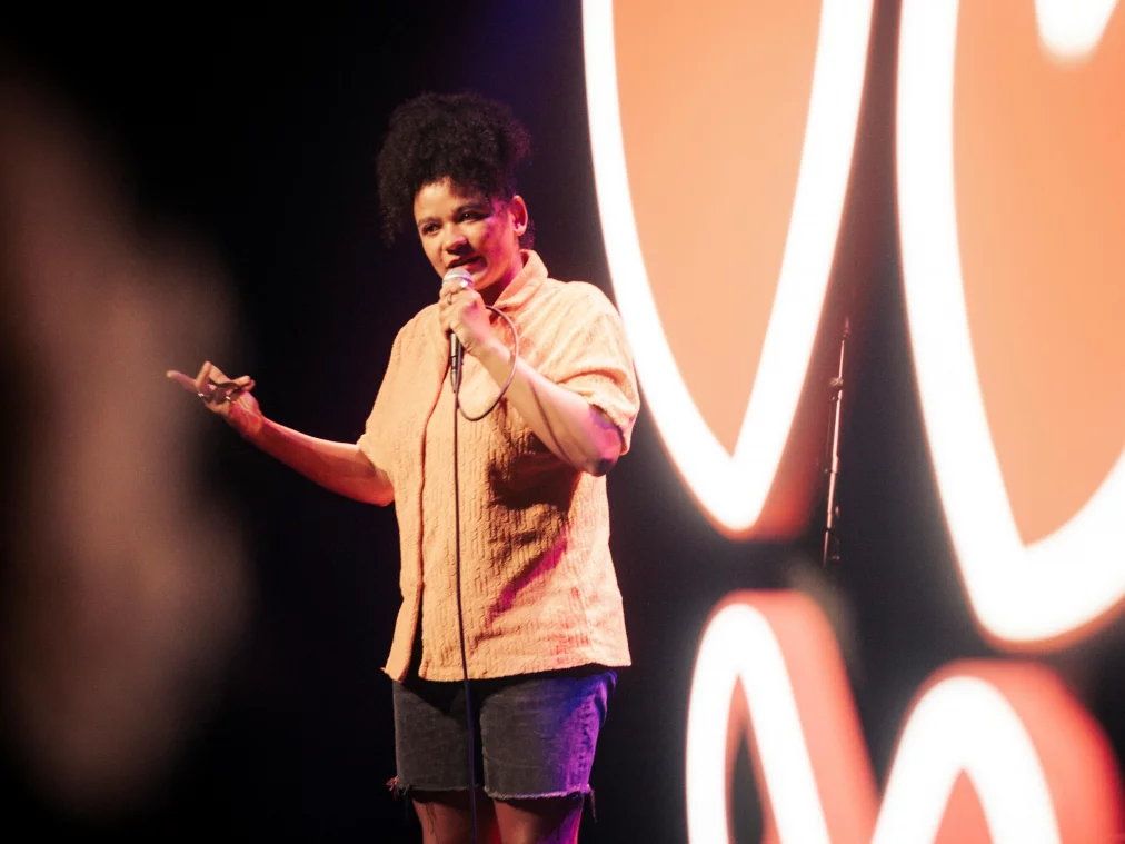 A person stands on a stage speaking into a handheld microphone. One hand is extended to the side, the body slightly leaning forward. Abstract, curved light shapes glow in the background against a dark stage setting.