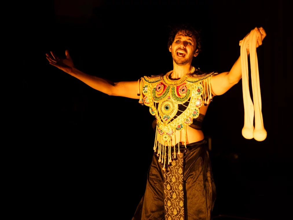 Eine Person trägt einen goldfarbenen, reich verzierten Brustschmuck und einen schwarzen Wickelrock. Mit ausgebreiteten Armen steht die Person im warmen Bühnenlicht und hält in einer Hand zwei verbundene Stoffbälle. Der Ausdruck wirkt erzählerisch und präsent.