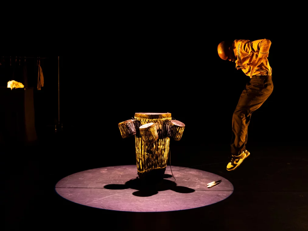 Eine Person im goldgelben Hemd springt in einer Lichtinsel in die Luft, der Körper nach vorn geneigt und die Beine angewinkelt. Unter der Person steht die trommelartige Skulptur, daneben liegen zwei kurze Stöcke. Der Rest der Bühne verschwindet im Schwarz.