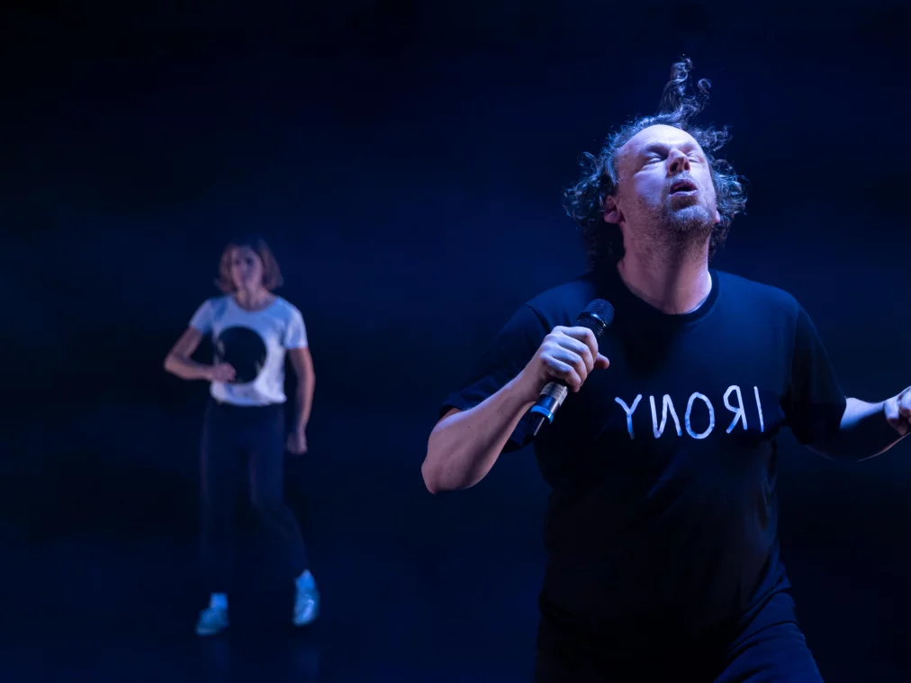 Eine Person mit Mikrofon steht im blauen Bühnennebel, der Kopf leicht zurückgeworfen und der Mund geöffnet, als würde die Person rufen oder singen. Im Hintergrund ist eine zweite Person in ähnlicher Bewegung verschwommen sichtbar.