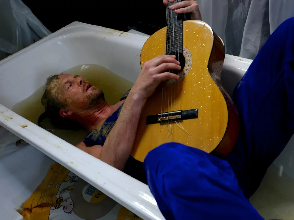 Eine Person liegt in einer mit Wasser gefüllten Badewanne und spielt eine Gitarre, während Arbeitskleidung und Requisiten die Szene umgeben.