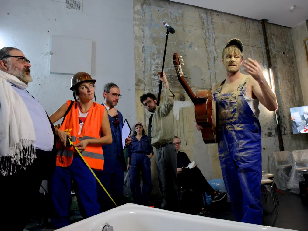 Eine Gruppe von Personen steht dicht beieinander in einem rauen Innenraum; eine Person mit Gitarre spricht gestikulierend, während andere zuhören.