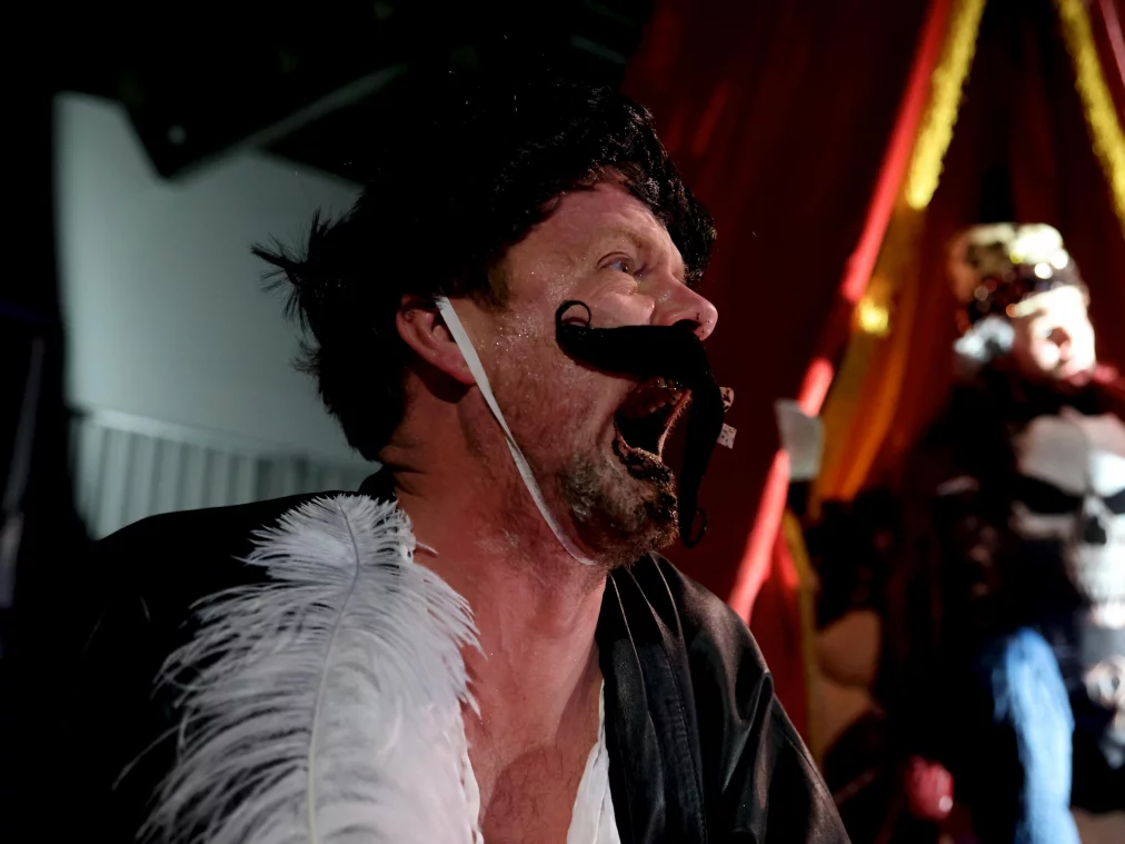 Nahaufnahme einer Person mit aufgesetztem falschem Schnurrbart, geöffnetem Mund und theatralischem Ausdruck vor rotem Bühnenlicht.