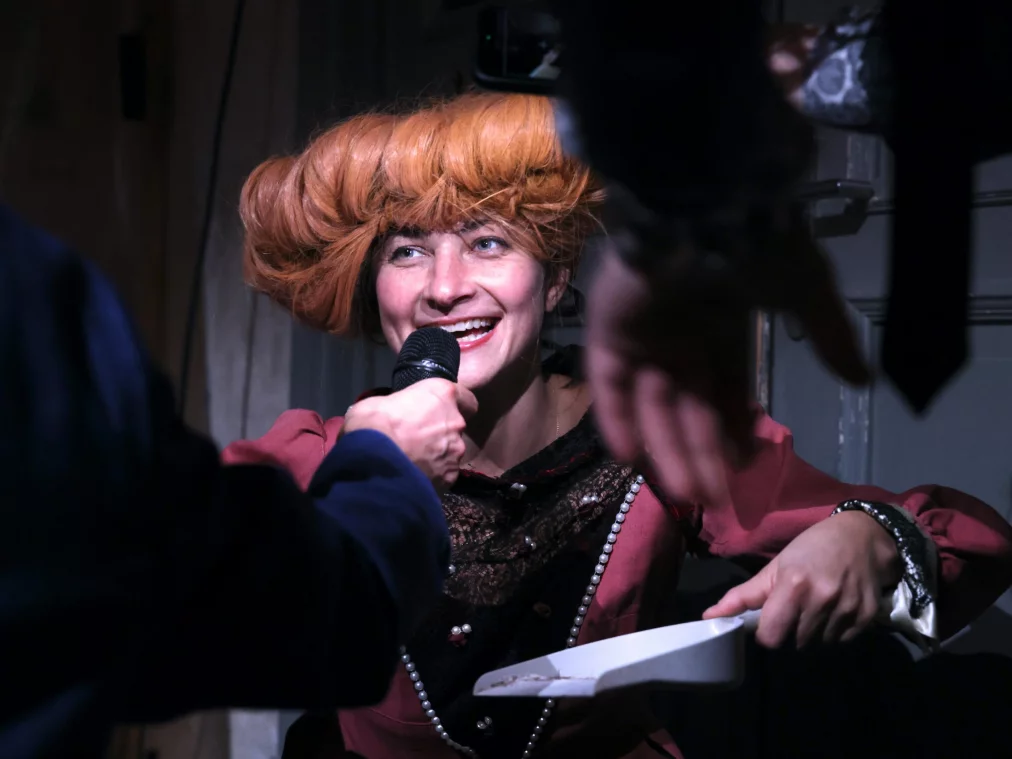 Eine lächelnde Person mit auffälliger, voluminöser Perücke spricht in ein Mikrofon, das ihr von einer anderen Person gehalten wird.