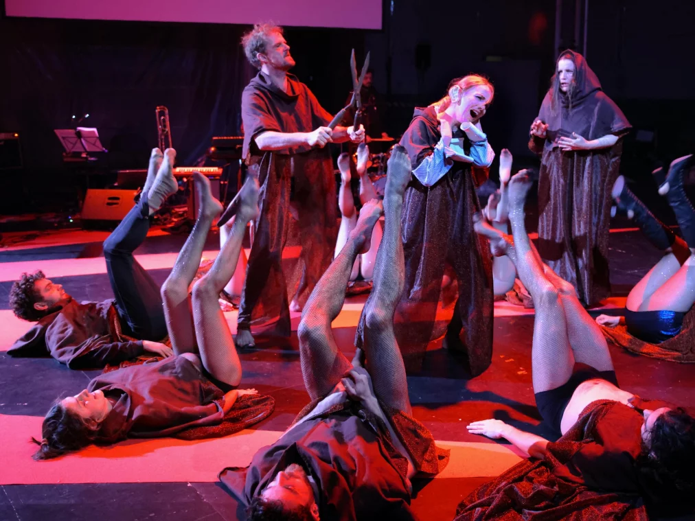 Eine Gruppe von Personen in dunklen Kostümen liegt auf dem Bühnenboden, die Beine angehoben, während drei stehende Personen mit ernsten Gesten in der Mitte agieren.