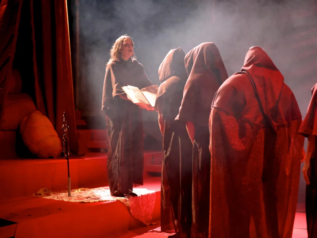 Mehrere Personen in dunklen, kapuzenartigen Gewändern stehen auf einer rot beleuchteten Bühne; eine Person hält ein geöffnetes Buch, aus dem Licht und Nebel aufsteigen.