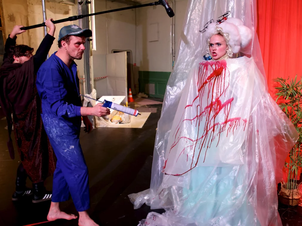 Eine Person in blauer Arbeitskleidung steht barfuß auf einer schwarzen Bühne und hält eine Kartuschenpistole in Richtung einer anderen Person. Die zweite Person steht regungslos in einem langen, hellen Kleid unter einer transparenten Kunststoffhülle, die mit roter Flüssigkeit bespritzt ist. Über der Szene hängt ein Mikrofon an einer Stange, im Hintergrund sind eine rote Vorhangfläche, eine Topfpflanze und technische Bühnenstrukturen zu sehen.