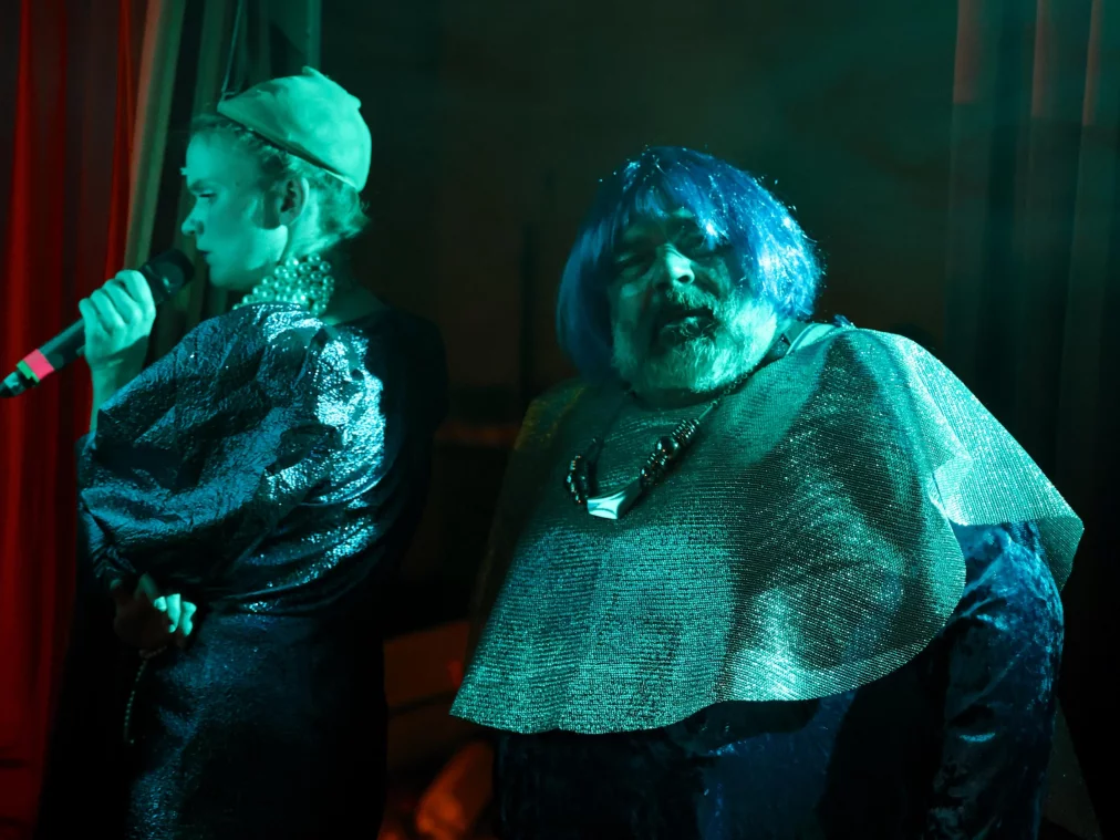 Zwei Personen stehen nebeneinander auf einer Bühne unter grünlichem Licht. Eine Person hält ein Mikrofon nahe am Mund und blickt zur Seite, während die andere Person frontal zur Kamera steht und ein glitzerndes Oberteil sowie eine auffällige Halskette trägt. Im Hintergrund hängen dunkle Vorhänge, die Szene wirkt wie ein musikalischer oder szenischer Auftritt.