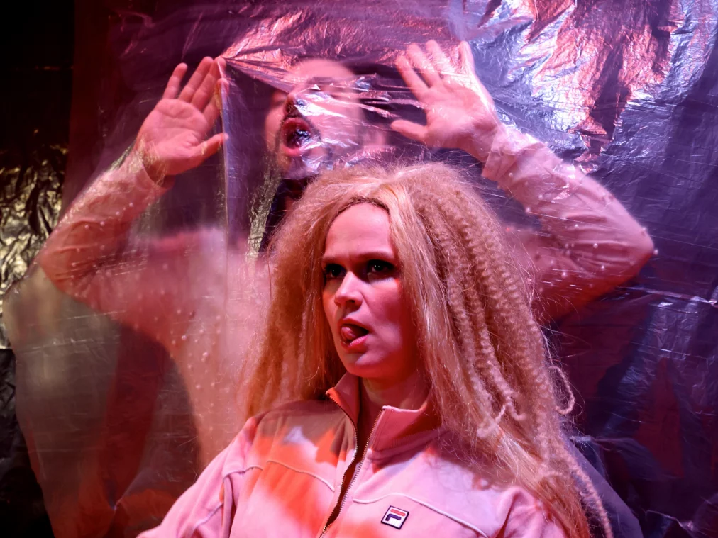 Eine Person mit offenem Mund drückt die Hände gegen eine transparente Folie, hinter der eine weitere Person schemenhaft sichtbar ist.