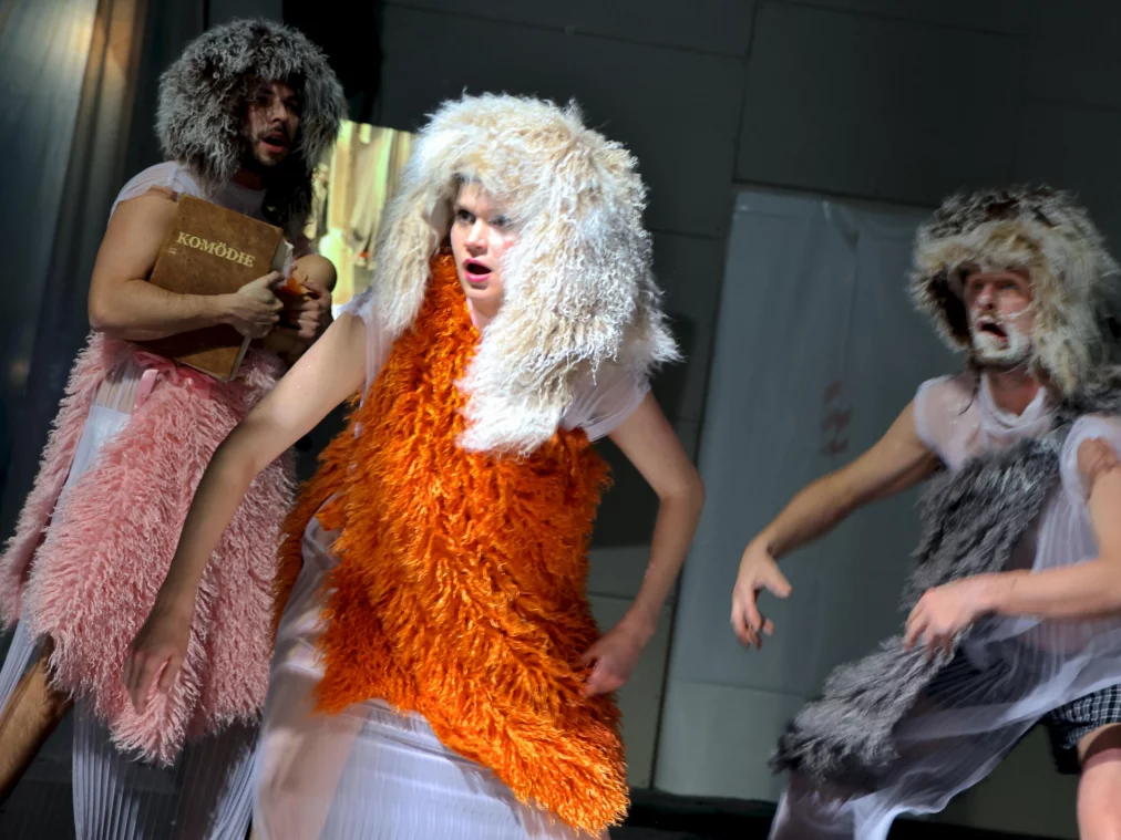 Drei Personen in zotteligen, tierähnlichen Kostümen bewegen sich mit ausdrucksstarken Gesten über eine helle Bühne.