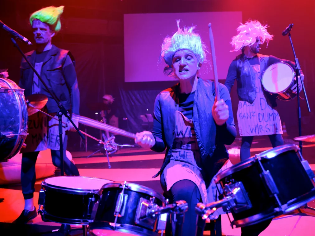 Mehrere Personen befinden sich auf einer Bühne mit Schlaginstrumenten. In der Mitte sitzt eine Person an einem Drumset und hebt zwei Schlägel, während weitere Personen mit Trommeln im Stehen oder Gehen spielen. Alle tragen dunkle Kleidung mit hellen Schürzen oder Schildern. Die Bühne ist in rotes und violettes Licht getaucht, im Hintergrund sind Mikrofone und weitere Instrumente zu sehen.