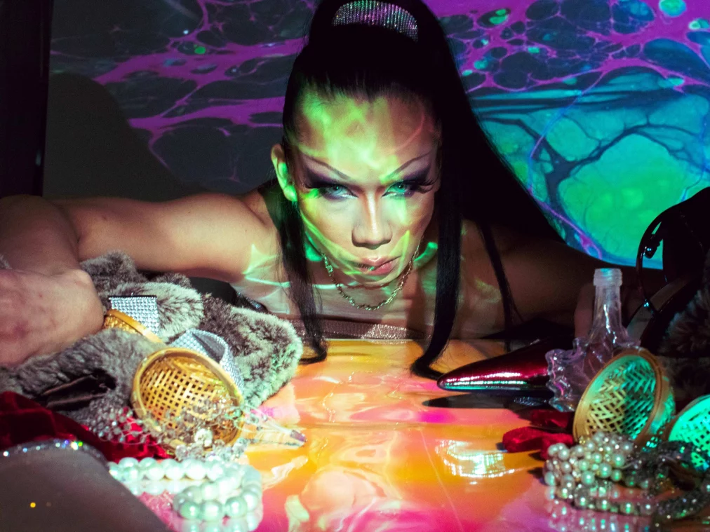 Eine Person liegt nach vorne gelehnt auf einer glänzenden, farbig reflektierenden Oberfläche und blickt intensiv direkt in die Kamera. Bunte Projektionen in Grün, Türkis und Violett überziehen Gesicht, Oberkörper und den Hintergrund. Um die Person herum liegen dekorative Objekte wie Perlenketten, Stoffe, Accessoires und glänzende Schuhe, die das Licht spiegeln und eine dichte, theatralische Atmosphäre erzeugen.