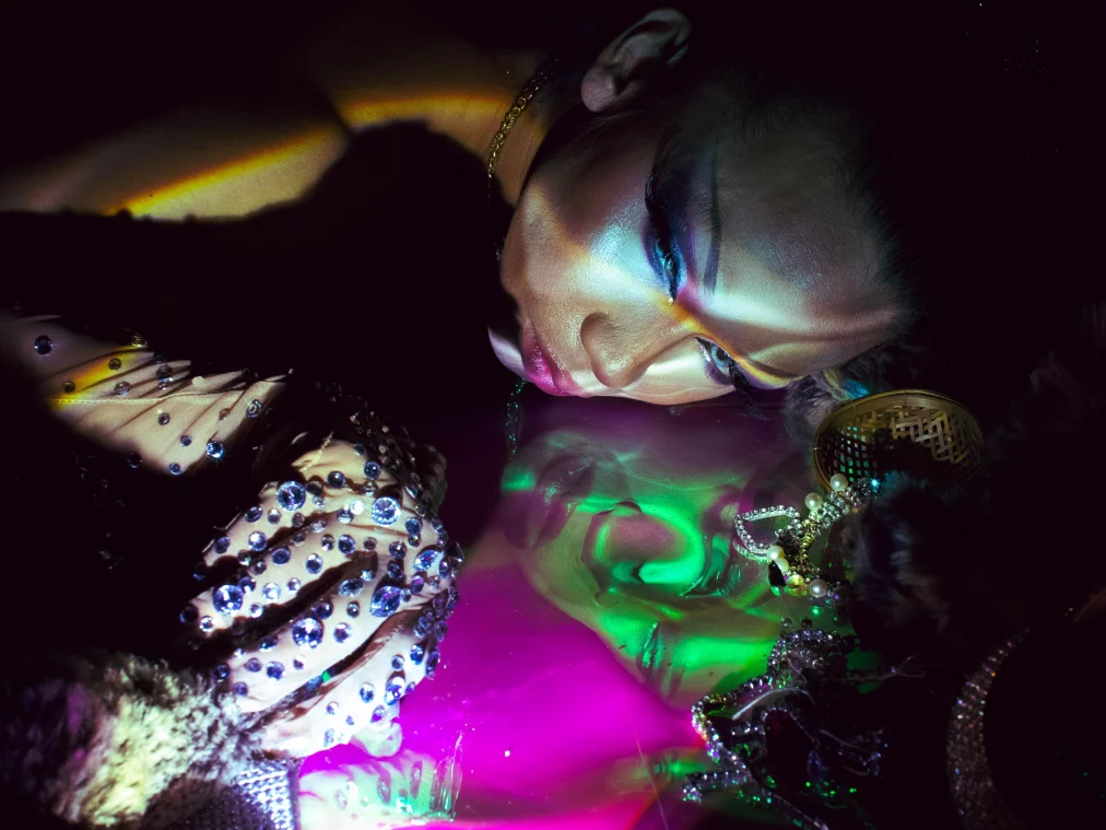 Eine Person liegt seitlich auf einer spiegelnden Fläche, das Gesicht von bunten Lichtprojektionen überzogen. Schmucksteine auf dem Handschuh funkeln, während sich das Gesicht klar in der glänzenden Oberfläche spiegelt.