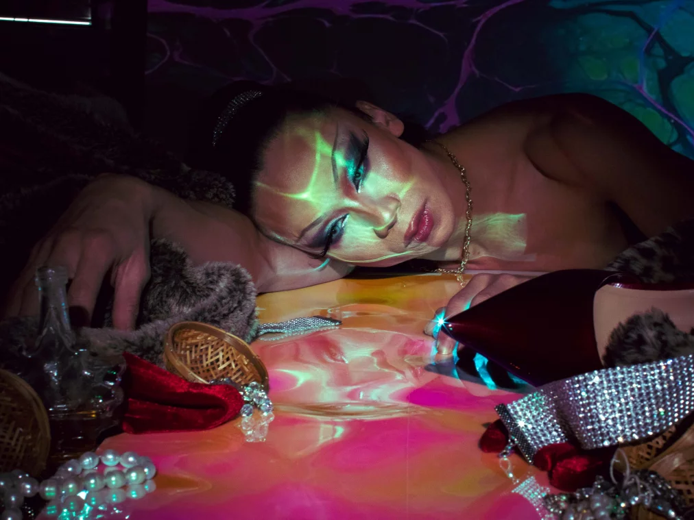 Eine Person liegt auf dem Unterarm gestützt inmitten von Pelzstoffen und glitzernden Gegenständen. Projektionen in Grün und Pink überziehen Gesicht und Umgebung, während die Spiegelung der Person in der glänzenden Fläche sichtbar wird.