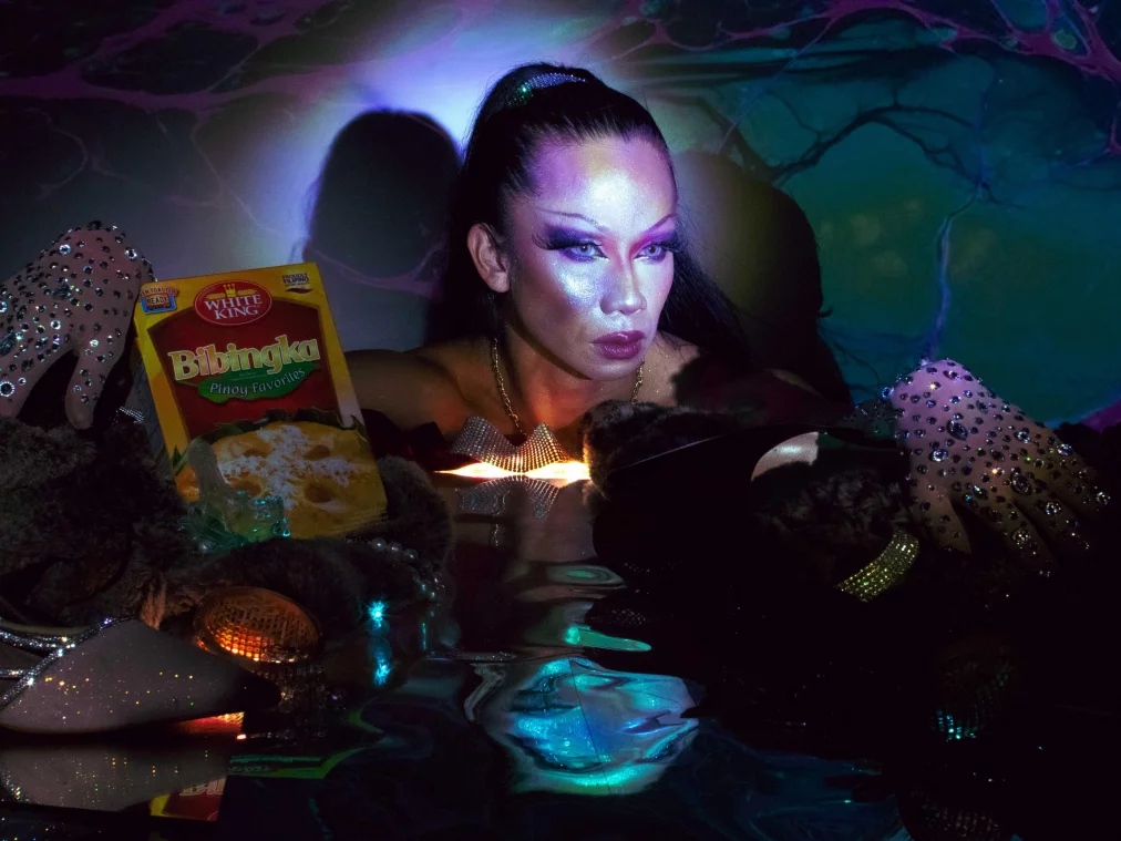 Eine Person mit starkem Make-up sitzt zwischen Accessoires, Glitzerhandschuhen und einer Verpackung für „Bibingka“. Projektionen in Blau und Violett beleuchten das Gesicht, das konzentriert nach vorn blickt, während sich die Farben im reflektierenden Untergrund brechen.