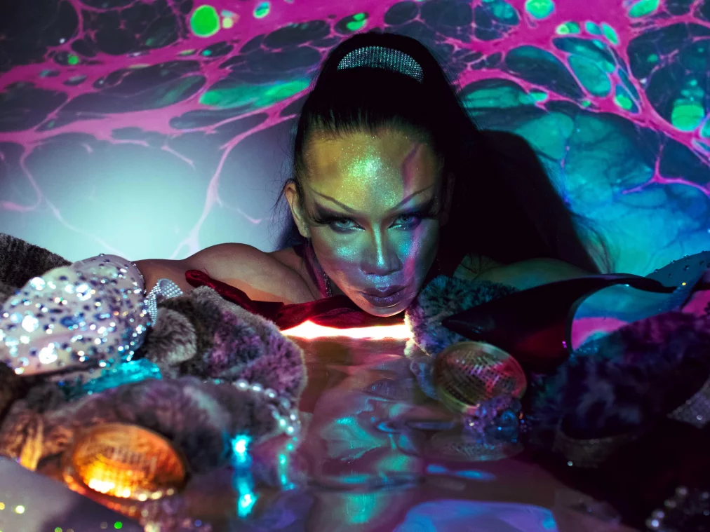 Eine Person mit stark geschminktem, glänzend schimmerndem Gesicht lehnt sich über einer spiegelnden Oberfläche, umgeben von Pelzstoffen, glitzernden Accessoires und Projektionen in Pink, Grün und Blau. Der Blick ist direkt und intensiv, die Lichtreflexe verzerren sich in der Spiegelung..