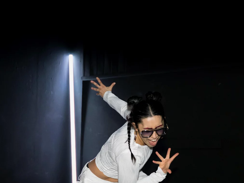 Eine Person kniet auf dem Boden und spannt den Körper dynamisch an. Ein Arm ist nach hinten oben geöffnet, der andere nach vorne gestreckt. Eine vertikale Lichtleiste leuchtet im Hintergrund, silberne Streifen liegen um die Person herum.