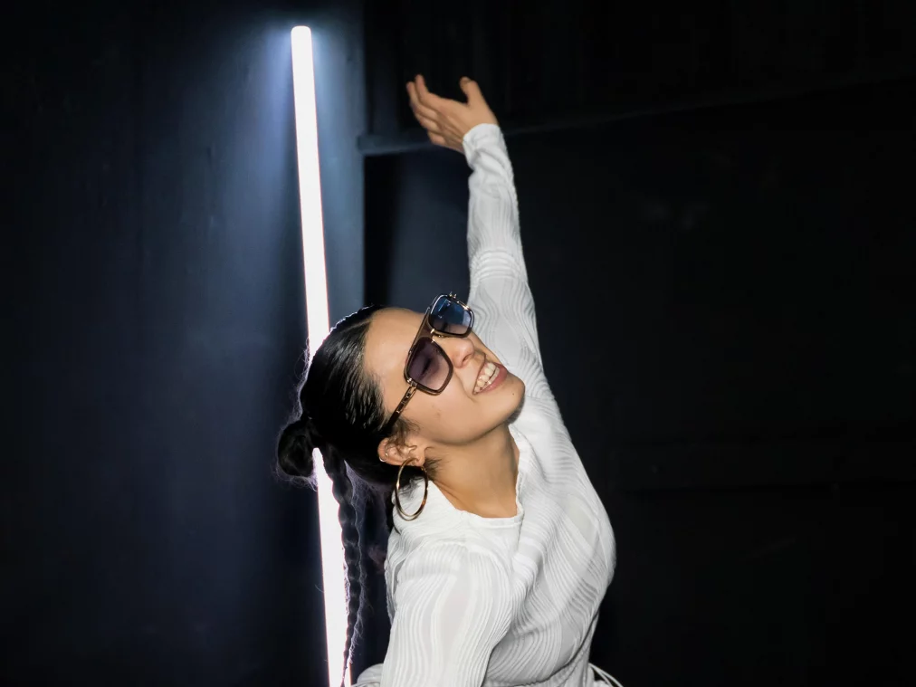 Eine Person kniet aufrecht auf dem Boden, stützt sich mit einer Hand ab und hebt den anderen Arm nach oben. Das Gesicht ist leicht nach oben gedreht. Hinter der Person steht eine helle vertikale Lichtleiste.