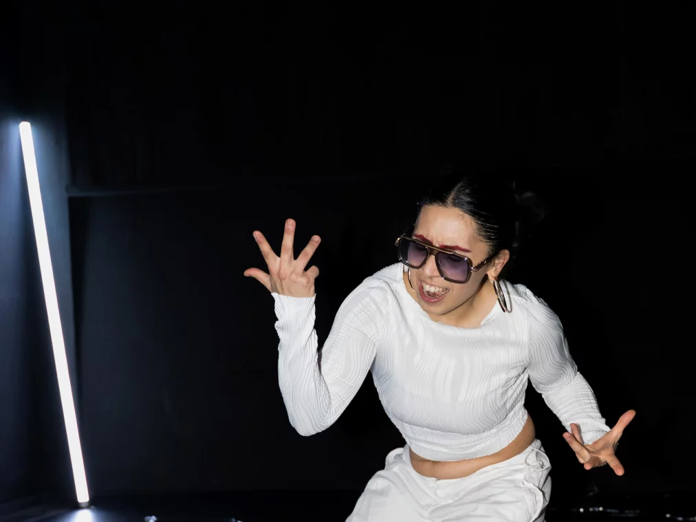 Eine Person kniet tief auf dem Boden und formt mit beiden Händen expressive Gesten. Der Oberkörper ist nach vorne geneigt, das Gesicht zeigt eine intensive Mimik. Eine vertikale Lichtleiste und silberne Bodenstreifen rahmen die Szene.