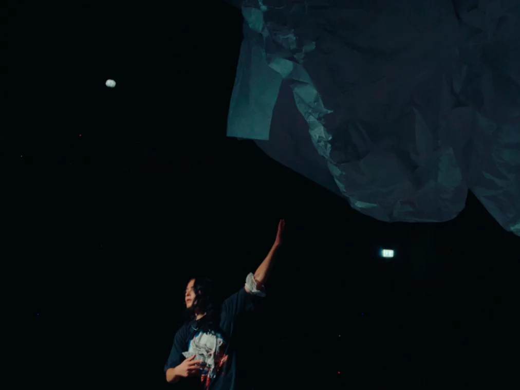 Eine einzel­ne Performer*in steht auf einer abgedunkelten Bühne, ein Arm ist nach oben gestreckt. Über ihr schwebt ein großes, zerknittertes, wolkenähnliches Tuch im Schein eines kühlen, blauen Lichts.