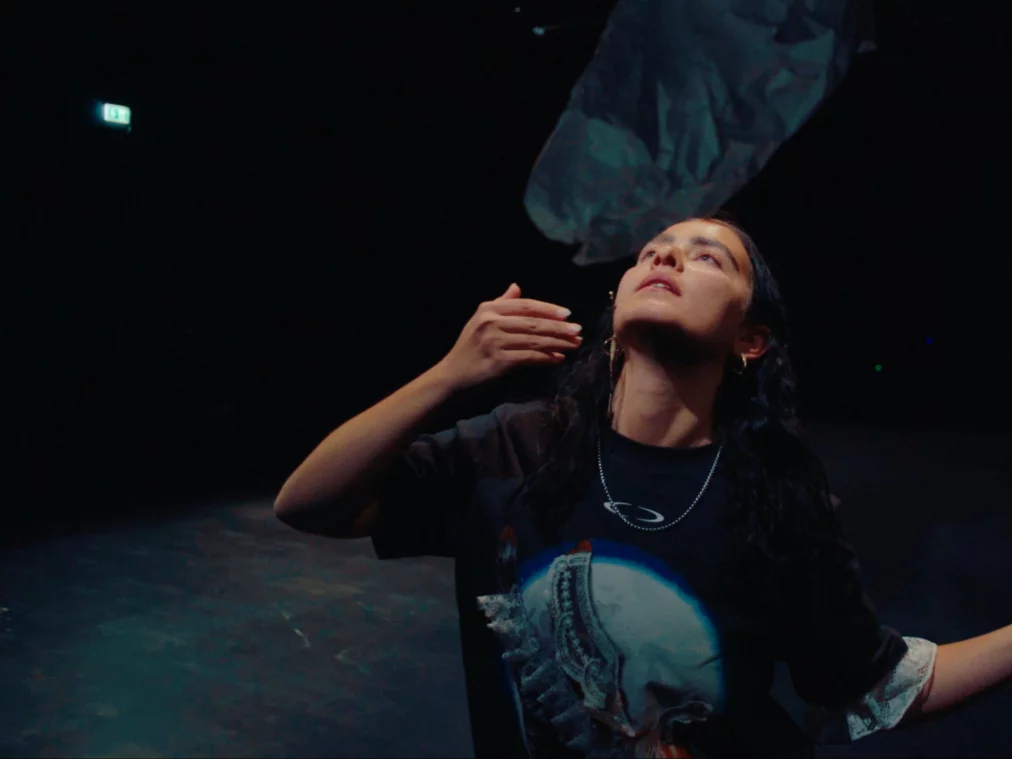 Eine Performer*in steht im Vordergrund und hebt den Blick suchend nach oben zum schwebenden, tuchartigen Objekt; ihre Arme sind leicht geöffnet in einer gestischen Bewegung.