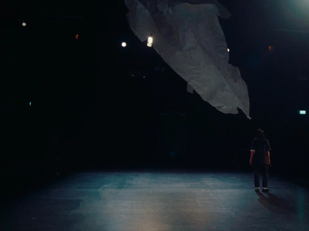 In einer fast leeren, dunklen Bühne geht eine Performer*in nach vorn, während ein riesiges, helles, papierartiges Tuch über ihr hängt und sich ausbreitet.