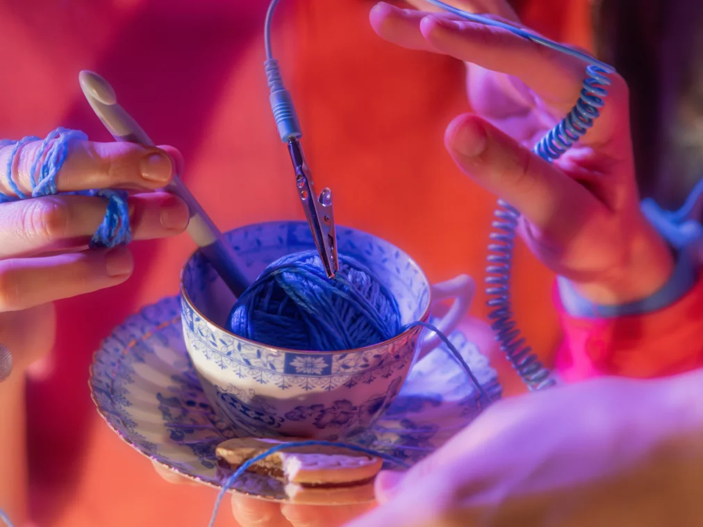 Eine Nahaufnahme zeigt mehrere Hände um eine blau-weiß gemusterte Teetasse mit blauer Wolle. Ein Häkelnadelgriff, eine Krokodilklemme und spiralförmige Kabel sind mit den Fäden und Händen verbunden.