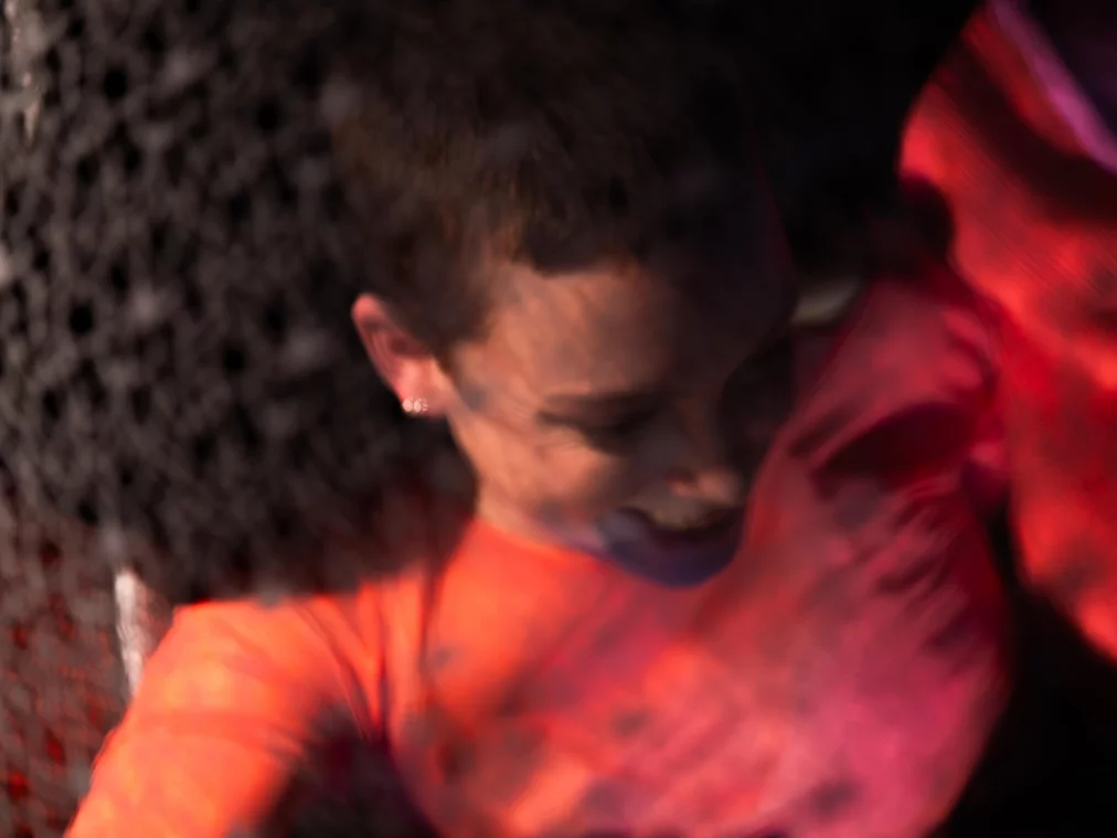 Eine Person in einem leuchtend roten Oberteil bewegt sich dynamisch vor einem dunklen, netzartigen Hintergrund. Das Bild ist unscharf und vermittelt eine starke Bewegung und körperliche Spannung.