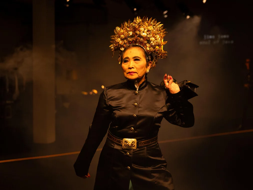Eine Person steht frontal auf der Bühne und trägt ein schwarzes Kostüm mit goldenen Details sowie eine große, goldene Kopfbedeckung. Warmes Licht fällt von oben, der Hintergrund ist dunkel und leicht verraucht.