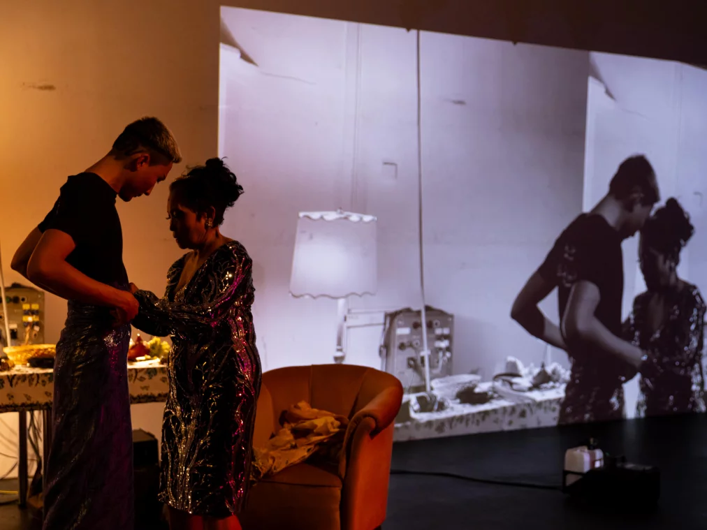 Zwei Personen stehen sich in einem Bühnenraum gegenüber. Eine Person hilft der anderen beim Anlegen eines glitzernden Kleidungsstücks. Im Hintergrund ist eine großformatige Schwarzweißprojektion derselben Situation zu sehen.