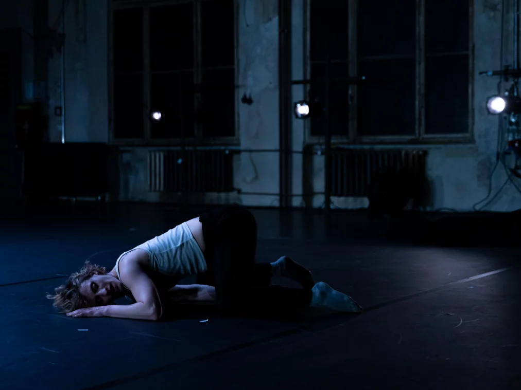 Eine Person liegt zusammengerollt auf dem Bühnenboden, der Kopf ruht auf den Armen, kühl blau beleuchtet.