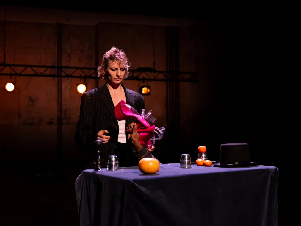 Eine Person steht hinter einem Tisch mit Requisiten, darunter Schuhe und Zitrusfrüchte, und blickt konzentriert nach unten.