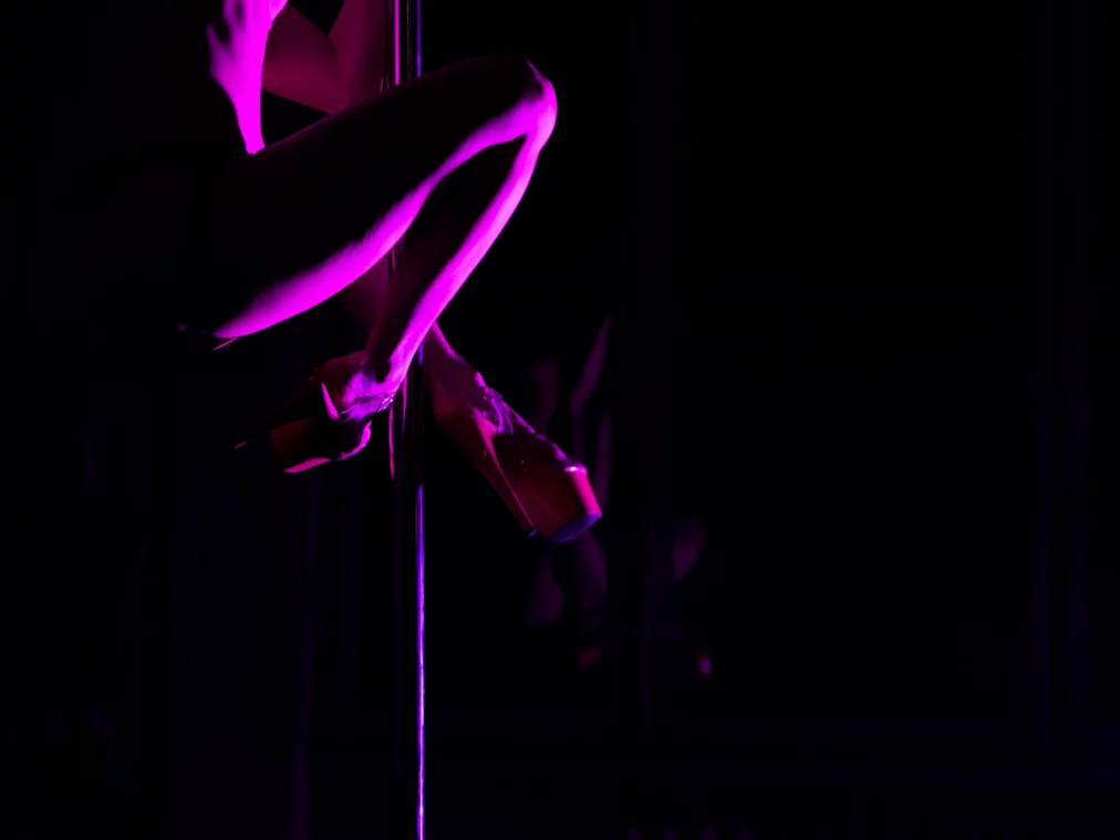 Nahaufnahme von Beinen in hohen Plateauschuhen, die eine Stange umschließen, violett beleuchtet vor dunklem Raum.