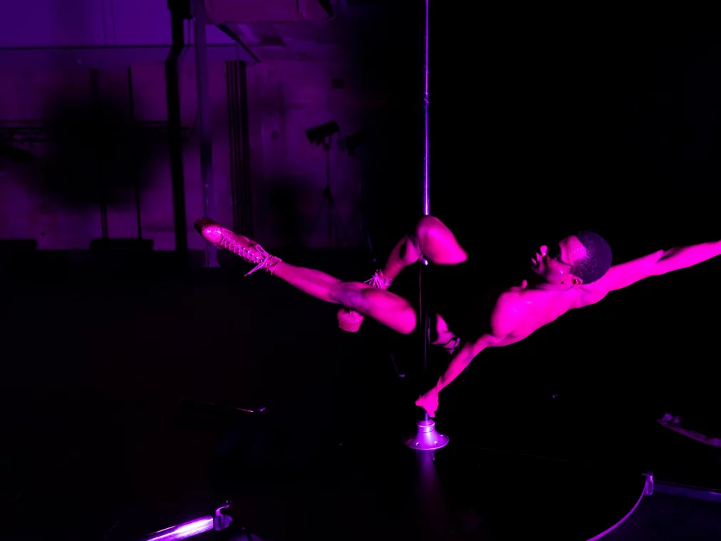 Eine Person hängt seitlich an einer Stange, der Körper horizontal ausgestreckt, Beine gespreizt, in violettem Bühnenlicht vor schwarzem Hintergrund.