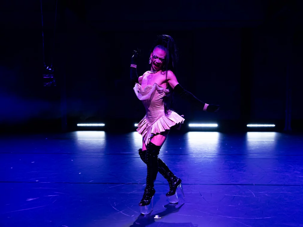 Eine Person in einem hellen Korsettkleid und hohen Plateauschuhen tanzt auf einer dunklen Bühne. Blaues Licht beleuchtet den Boden, während die Person mit angewinkelten Armen und leicht gedrehter Körperhaltung eine elegante Bewegung ausführt.