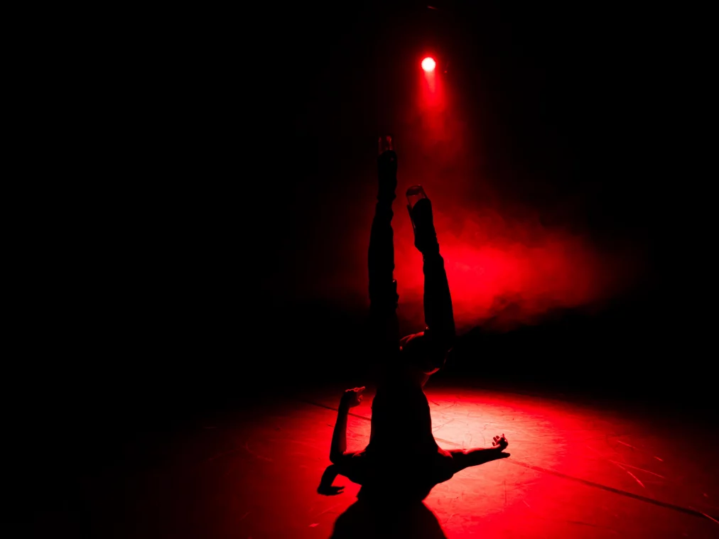 Eine Person befindet sich kopfüber auf dem Boden der Bühne, die Beine senkrecht nach oben gestreckt. Rotes Licht und Nebel erzeugen eine intensive, körperbetonte Szene.
