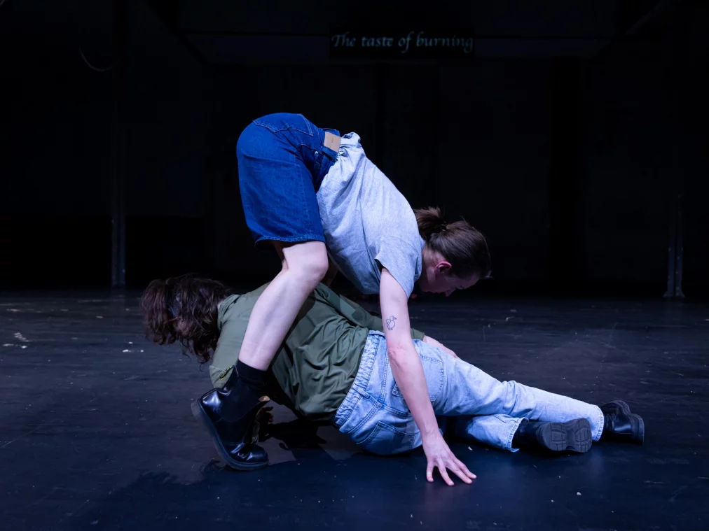 Zwei Personen befinden sich bodennah auf einer dunklen Bühne. Eine Person liegt auf dem Rücken, während eine andere darüber positioniert ist und das Gewicht verlagert. Die Körper bilden eine verschränkte, instabile Konstellation.+IsabellaFernandesSantana©MayraWallraff-29