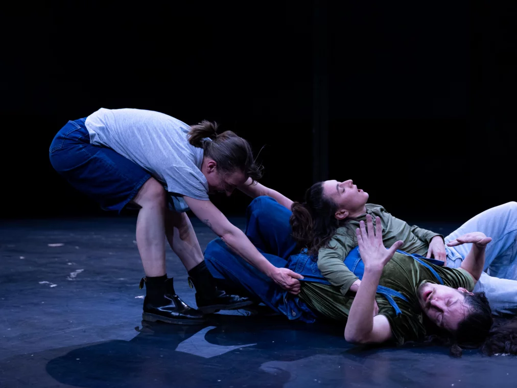 Zwei Personen liegen auf dem Boden, während eine dritte Person sich darüber beugt und an der Kleidung einer der liegenden Personen zieht. Die Körper stehen in enger, spannungsvoller Beziehung zueinander.