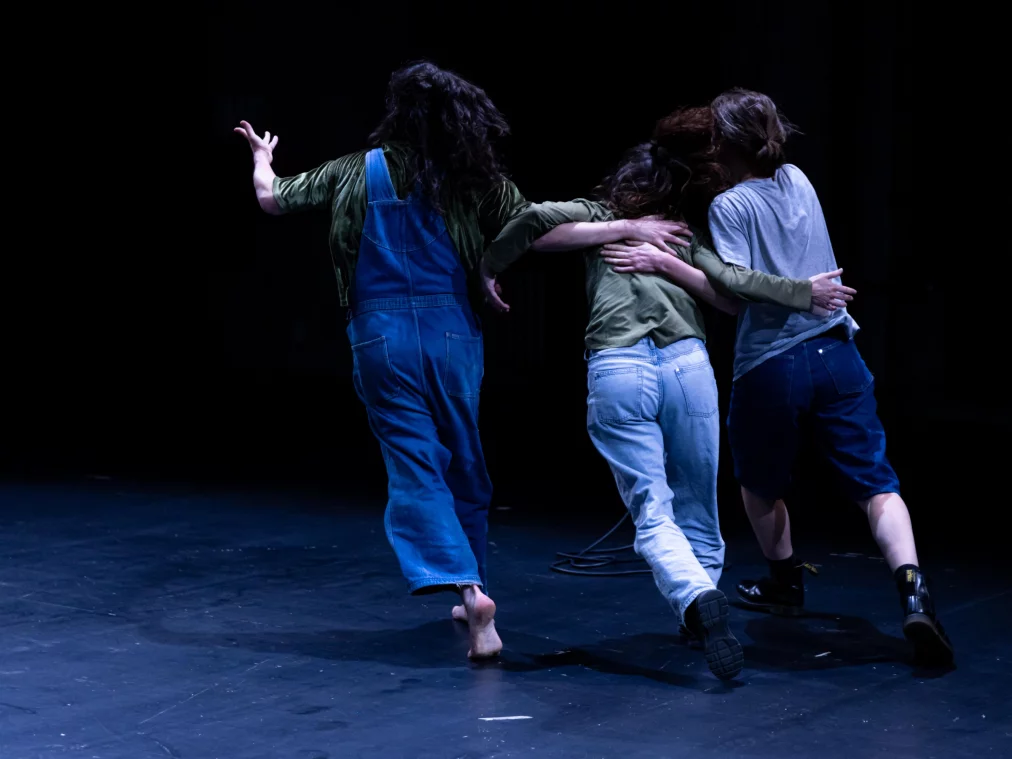 Drei Personen bewegen sich eng verbunden über die Bühne. Zwei Personen stützen eine dritte Person seitlich, während sie gemeinsam nach vorne gehen. Die Körperhaltung vermittelt Unterstützung und kollektive Bewegung.
