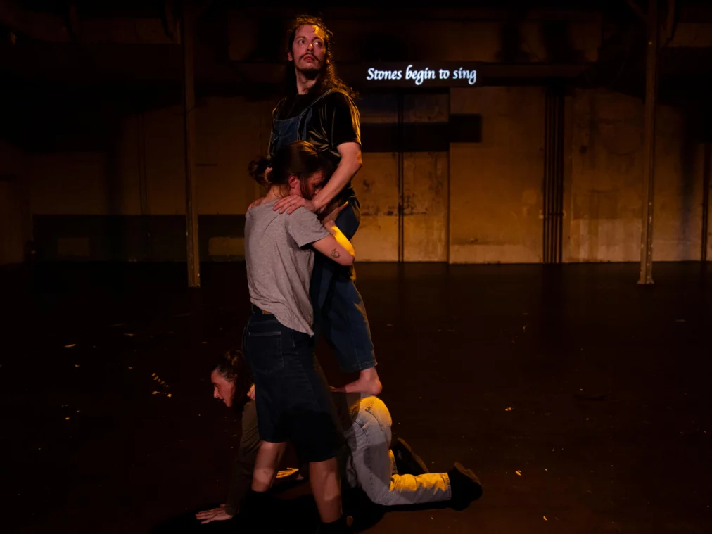 Drei Personen bilden eine vertikale Körperkonstruktion auf einer Bühne. Eine Person steht erhöht, während zwei Personen darunter kniend und gestützt eine Basis bilden. Im Hintergrund ist projizierter Text an der Wand sichtbar.