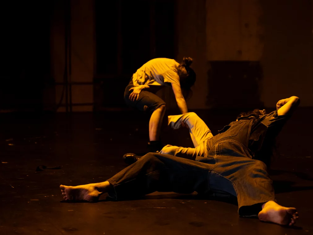 Mehrere Personen befinden sich in bodennahen Positionen auf einer dunkel beleuchteten Bühne. Eine Person liegt ausgestreckt, während eine andere sich darüber beugt. Warmes, gelbliches Licht erzeugt eine intensive, körperliche Atmosphäre.