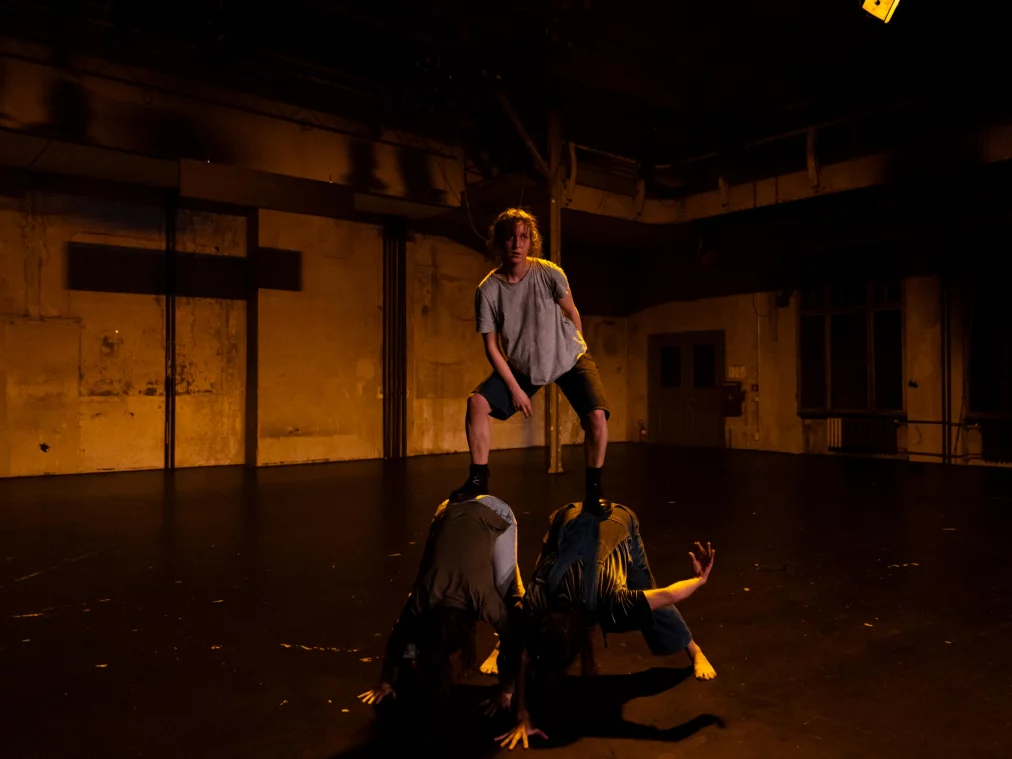 Eine Person steht mit gespreizten Beinen erhöht auf zwei anderen Personen, die sich nach unten abstützen. Der Raum wirkt industriell und leer, das Licht ist warm und konzentriert auf die Körper.