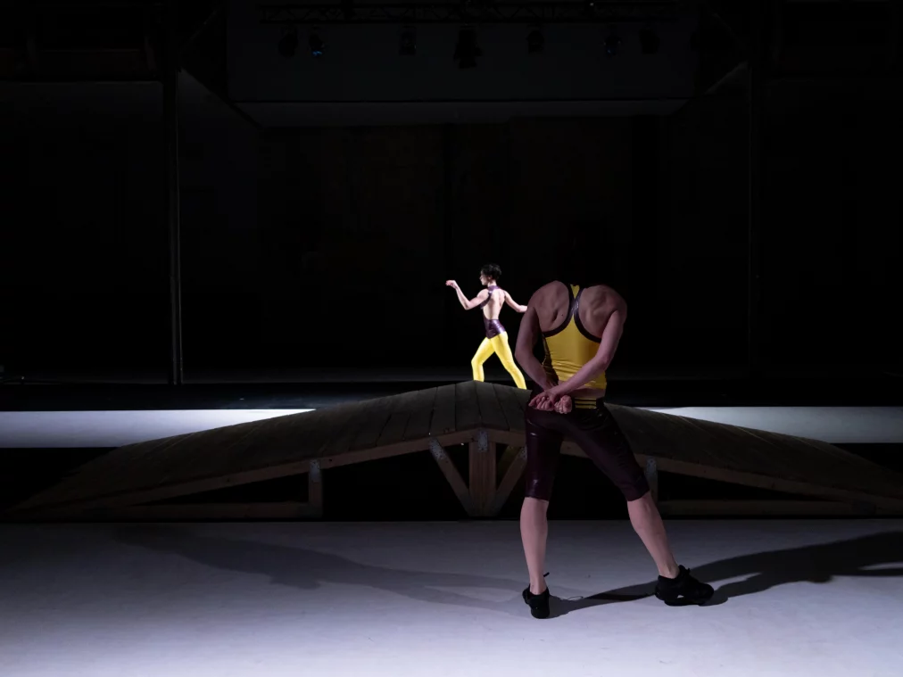 Zwei Personen stehen sich auf einer breiten Bühne gegenüber, getrennt durch einen dunklen Mittelstreifen. Beide Körper sind in unterschiedliche Richtungen ausgerichtet, die Bewegungen wirken kontrolliert und distanziert.